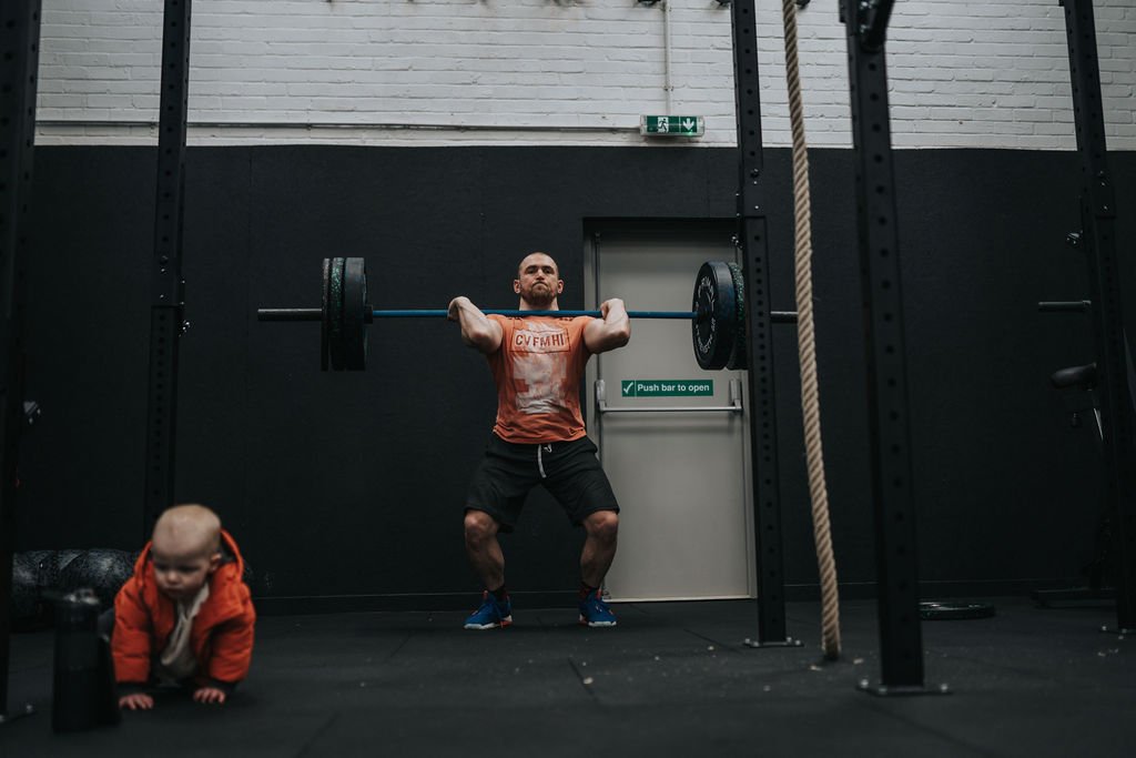 Man lifting weights in gym with baby nearby.